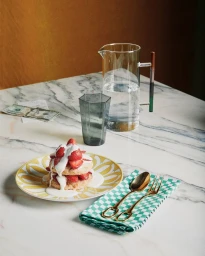 A plate of strawberry shortcake with cream, a glass pitcher of water, a glass, and gold cutlery on a marble table.
