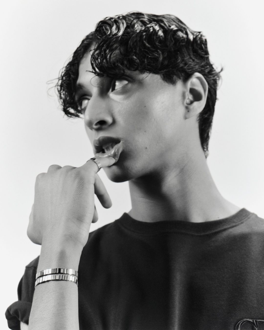 Monochrome portrait of a young man with curly hair, wearing a dark shirt and silver bracelets, posing in a studio environment.