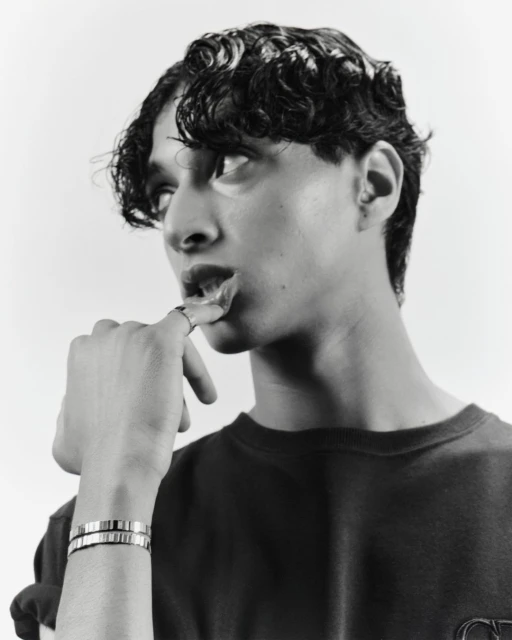 Monochrome portrait of a young man with curly hair, wearing a dark shirt and silver bracelets, posing in a studio environment.