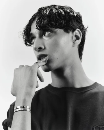 Monochrome portrait of a young man with curly hair, wearing a dark shirt and silver bracelets, posing in a studio environment.