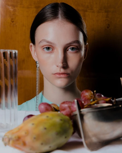 Close-up portrait of a woman with elegant earrings, paired with vibrant fruits and a reflective silver bowl on a white surface.