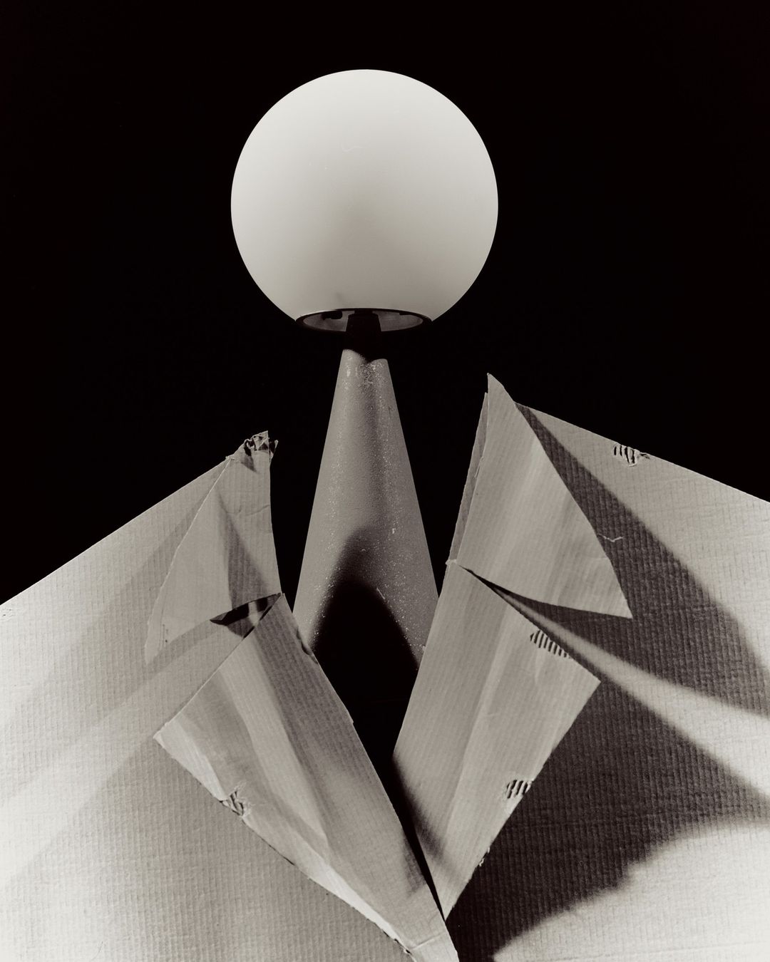 Abstract still life of an illuminated sphere atop a conical stand, with a paper jacket forming lapels, against a dark background.