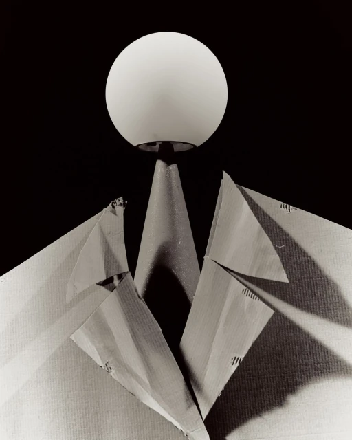 Abstract still life of an illuminated sphere atop a conical stand, with a paper jacket forming lapels, against a dark background.