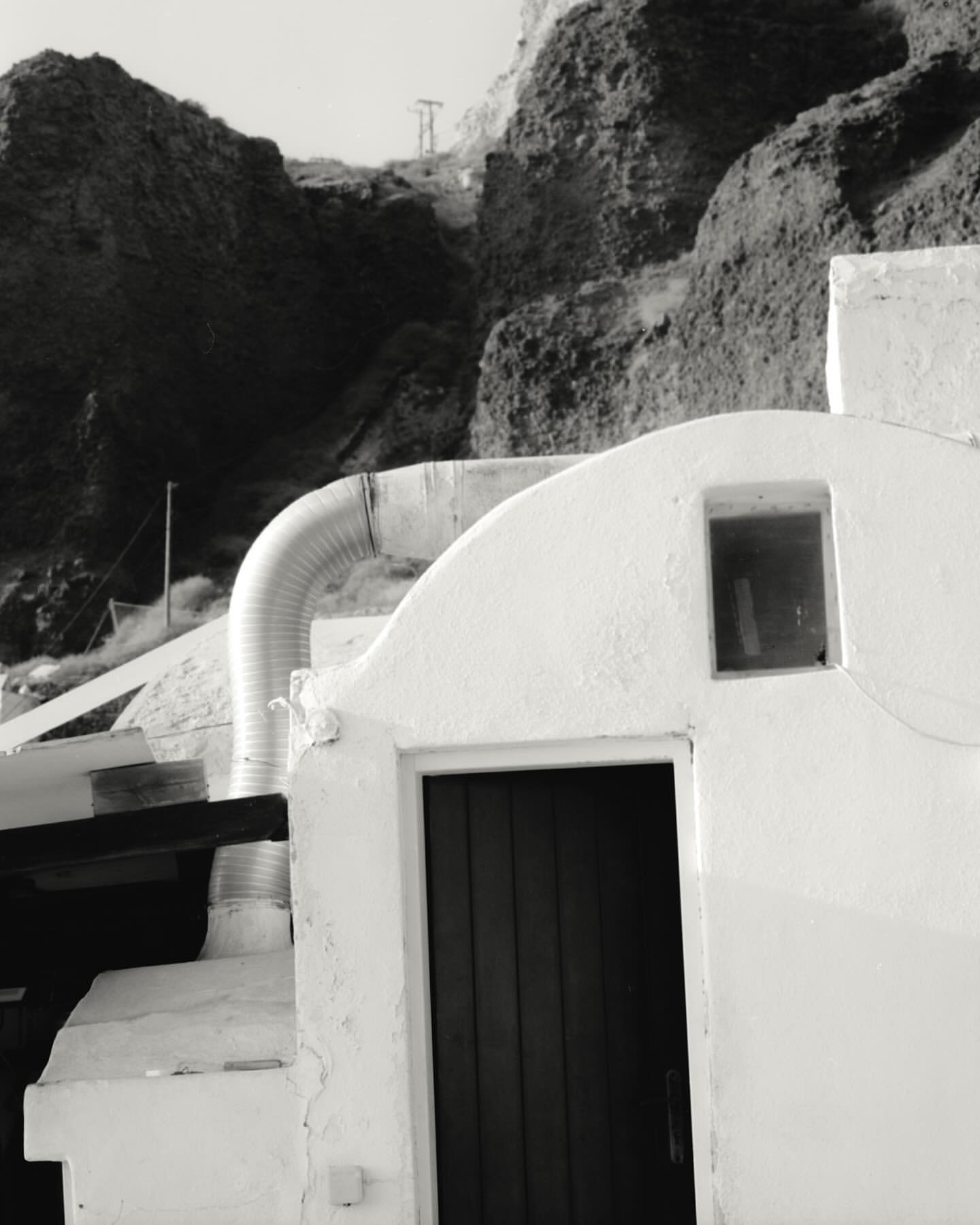 Monochrome architectural shot of a white building with a dark doorway, set against a rocky hillside. The style is minimalist.