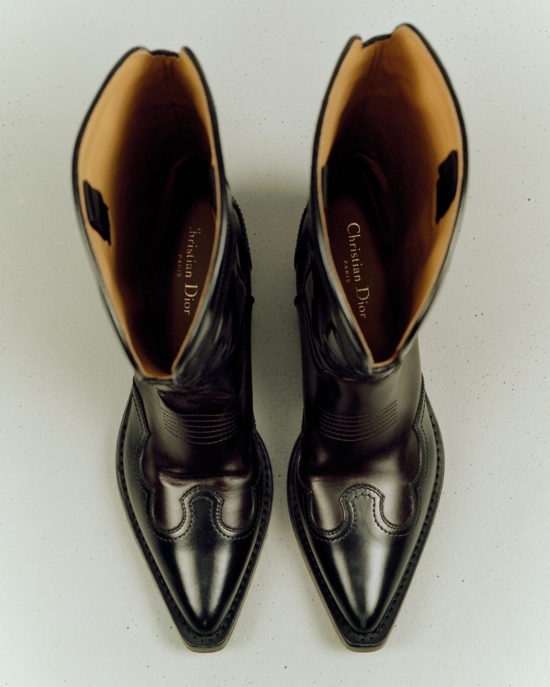 High-angle, close-up studio shot of a pair of black leather cowboy boots with light brown lining. The logo of Christian Dior is visible.