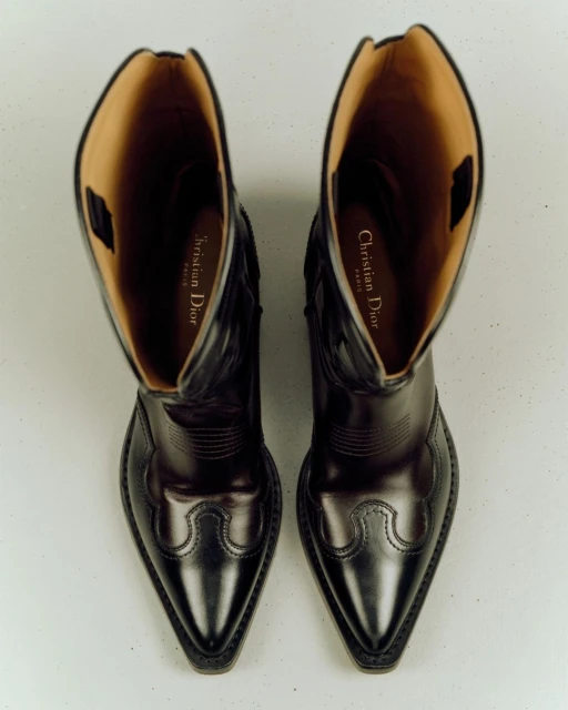 High-angle, close-up studio shot of a pair of black leather cowboy boots with light brown lining. The logo of Christian Dior is visible.