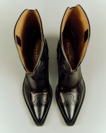 High-angle, close-up studio shot of a pair of black leather cowboy boots with light brown lining. The logo of Christian Dior is visible.