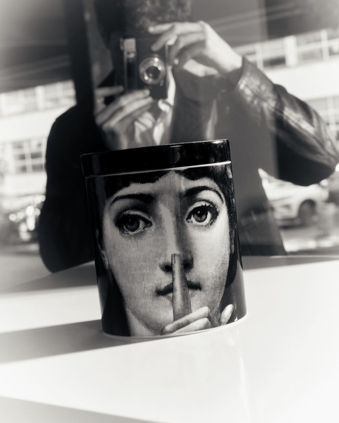 Black and white photograph featuring a decorative container with a portrait and a blurry reflection of a man holding a camera.