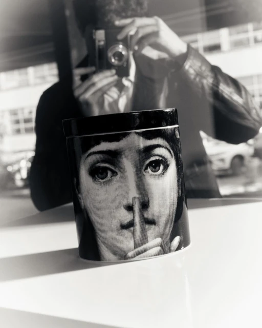 Black and white photograph featuring a decorative container with a portrait and a blurry reflection of a man holding a camera.
