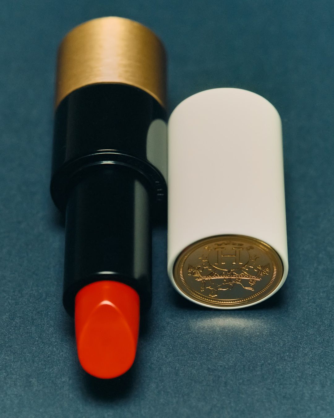 High-angle, close-up studio shot: red lipstick with gold accents next to a white cap revealing a gold coin on a blue background.