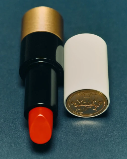High-angle, close-up studio shot: red lipstick with gold accents next to a white cap revealing a gold coin on a blue background.