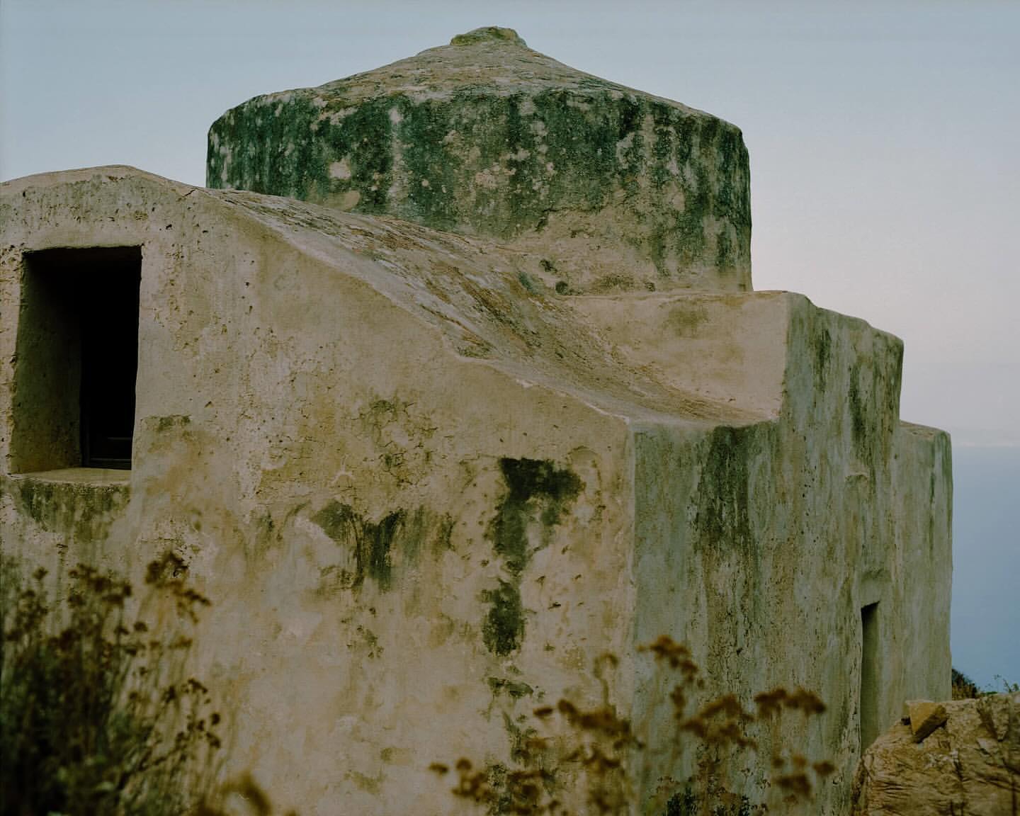Old, weathered stone building with a domed roof against a pale sky, showing architectural details and the texture of the building material.
