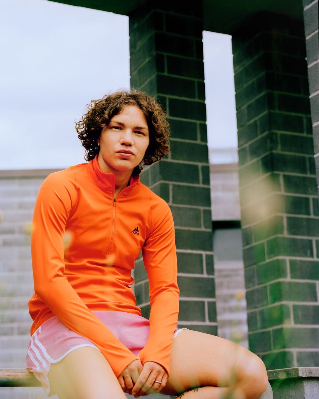 Athletic woman in an orange jacket sits outside a modern brick building; sports and lifestyle apparel visual content for advertising.