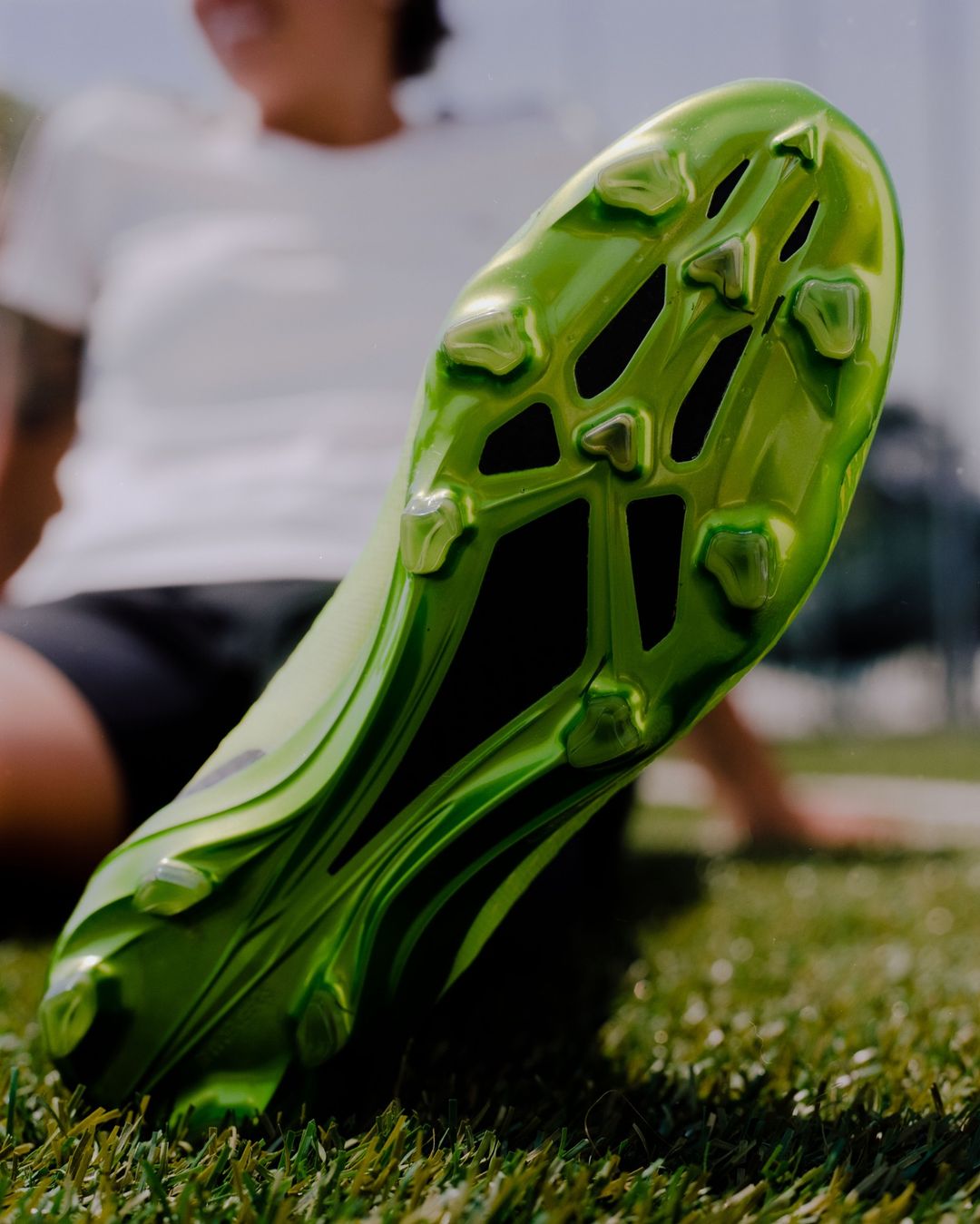 Close-up of a green soccer cleat on green grass with a person in a white shirt sitting in the blurred background.