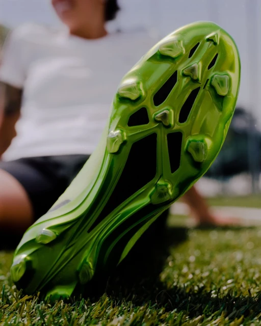 Close-up of a green soccer cleat on green grass with a person in a white shirt sitting in the blurred background.