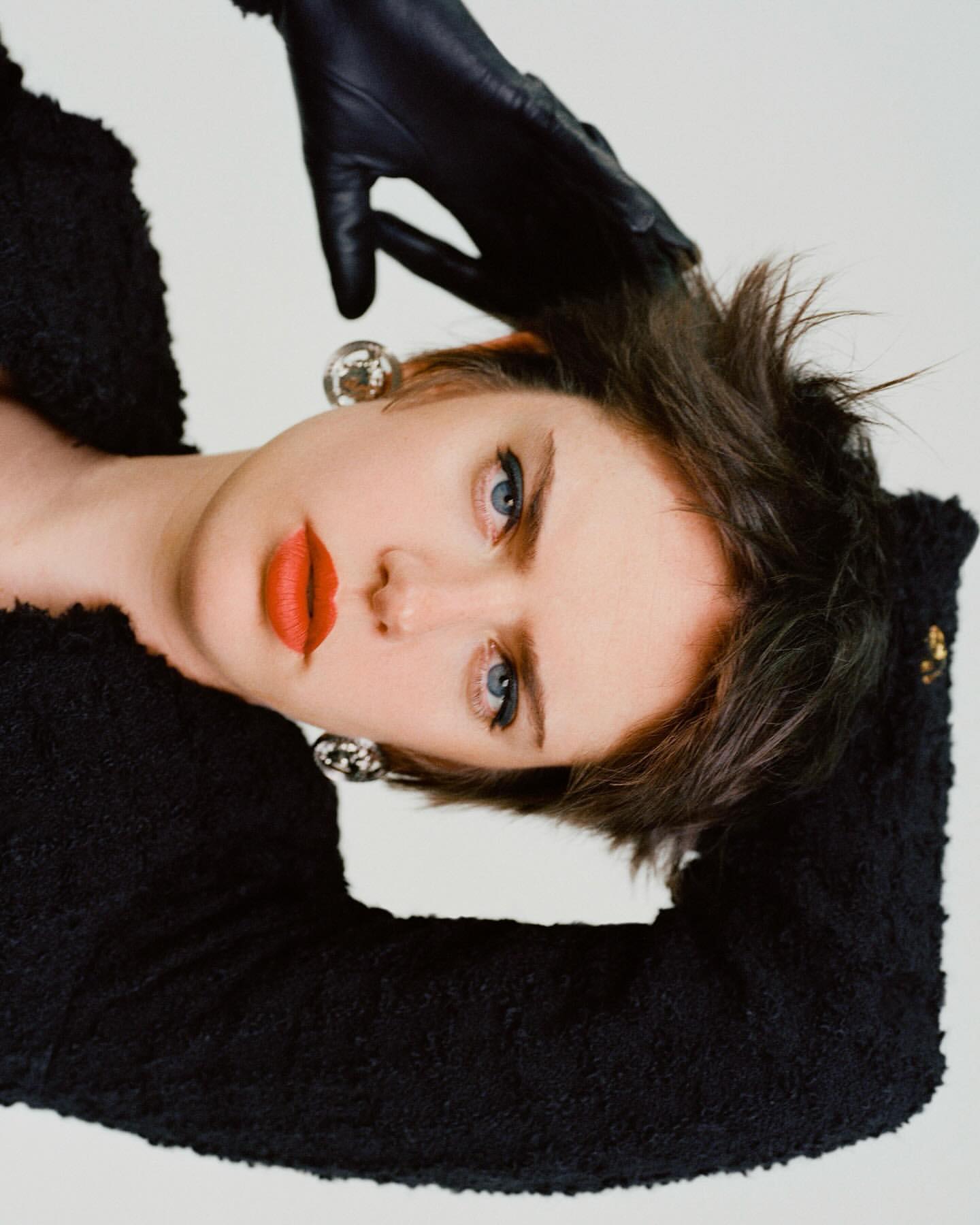 Portrait of a woman with short dark hair wearing a black textured jacket, red lipstick, leather gloves, and large silver earrings.