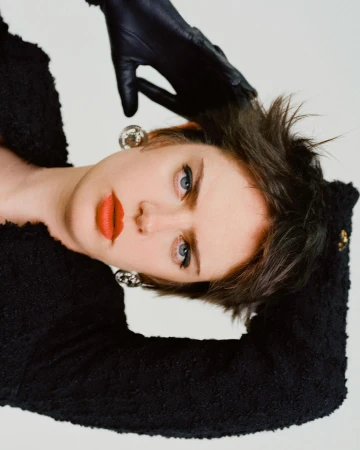 Portrait of a woman with short dark hair wearing a black textured jacket, red lipstick, leather gloves, and large silver earrings.