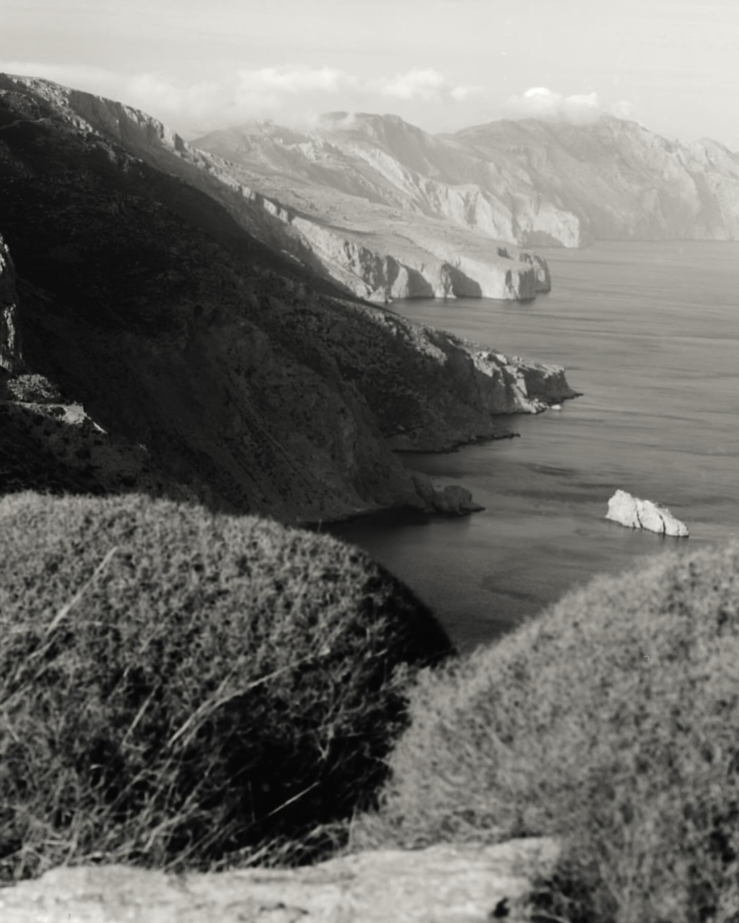 Black and white photograph of a rugged coastline with steep cliffs, a calm sea, and hazy mountains under an overcast sky.