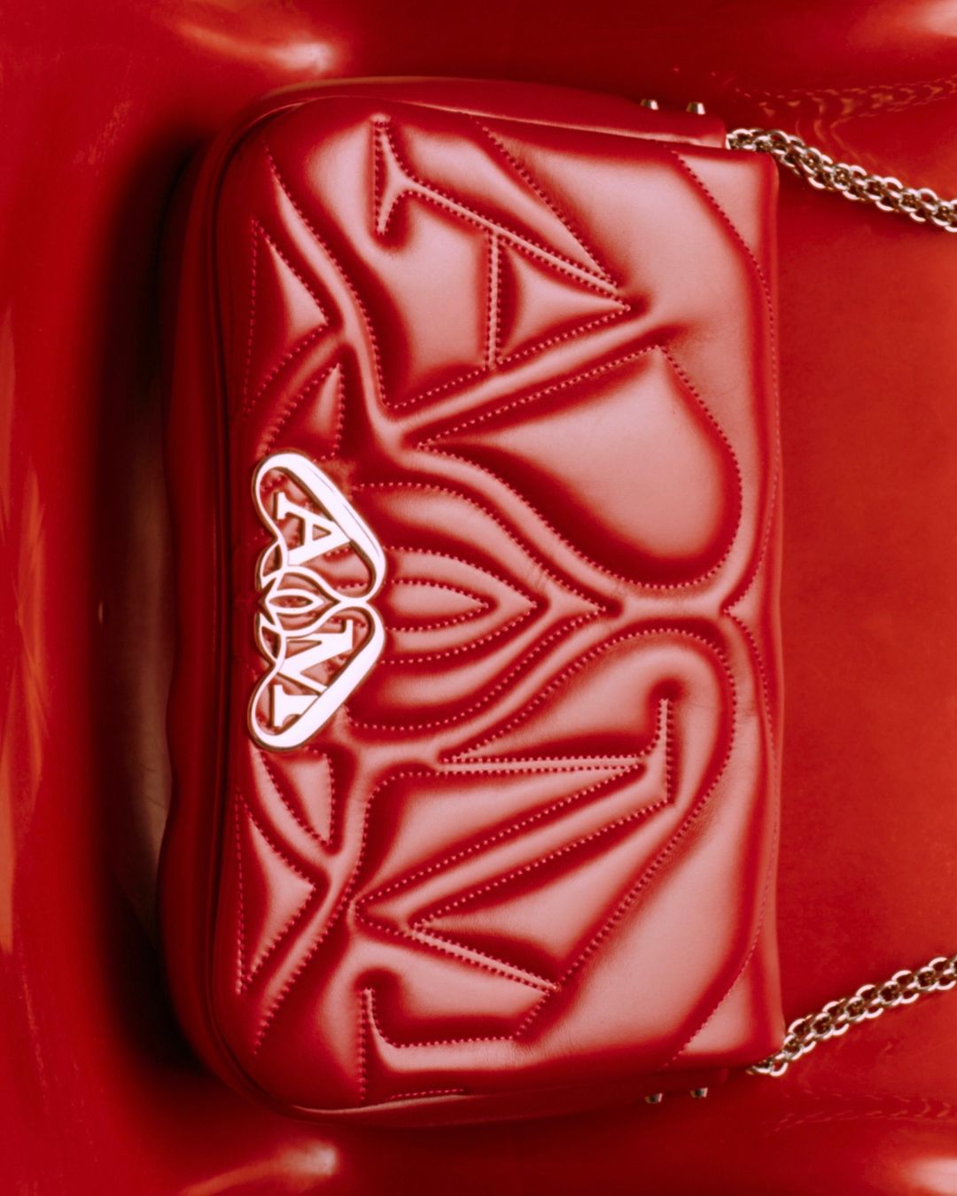 Red Alexander McQueen purse with chain strap on a glossy red surface, showcasing texture and a metal logo in a studio setting.