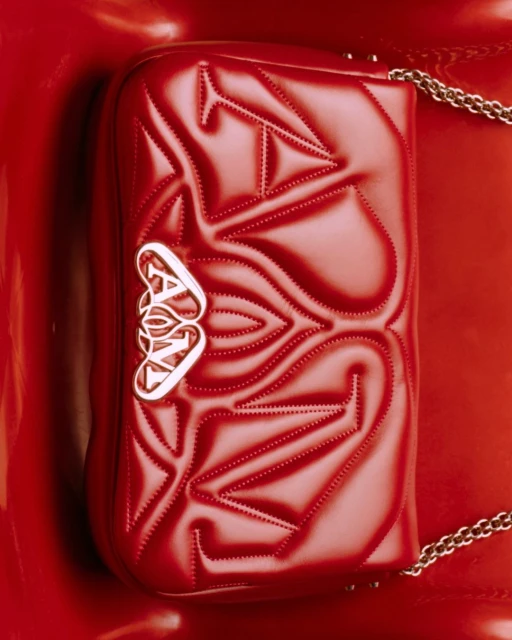 Red Alexander McQueen purse with chain strap on a glossy red surface, showcasing texture and a metal logo in a studio setting.