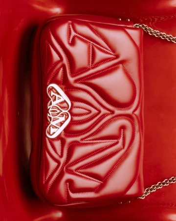 Red Alexander McQueen purse with chain strap on a glossy red surface, showcasing texture and a metal logo in a studio setting.