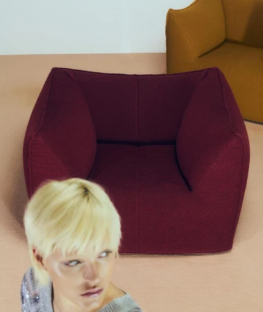 Blonde woman looking away in foreground with a dark red armchair and gold armchair in interior setting