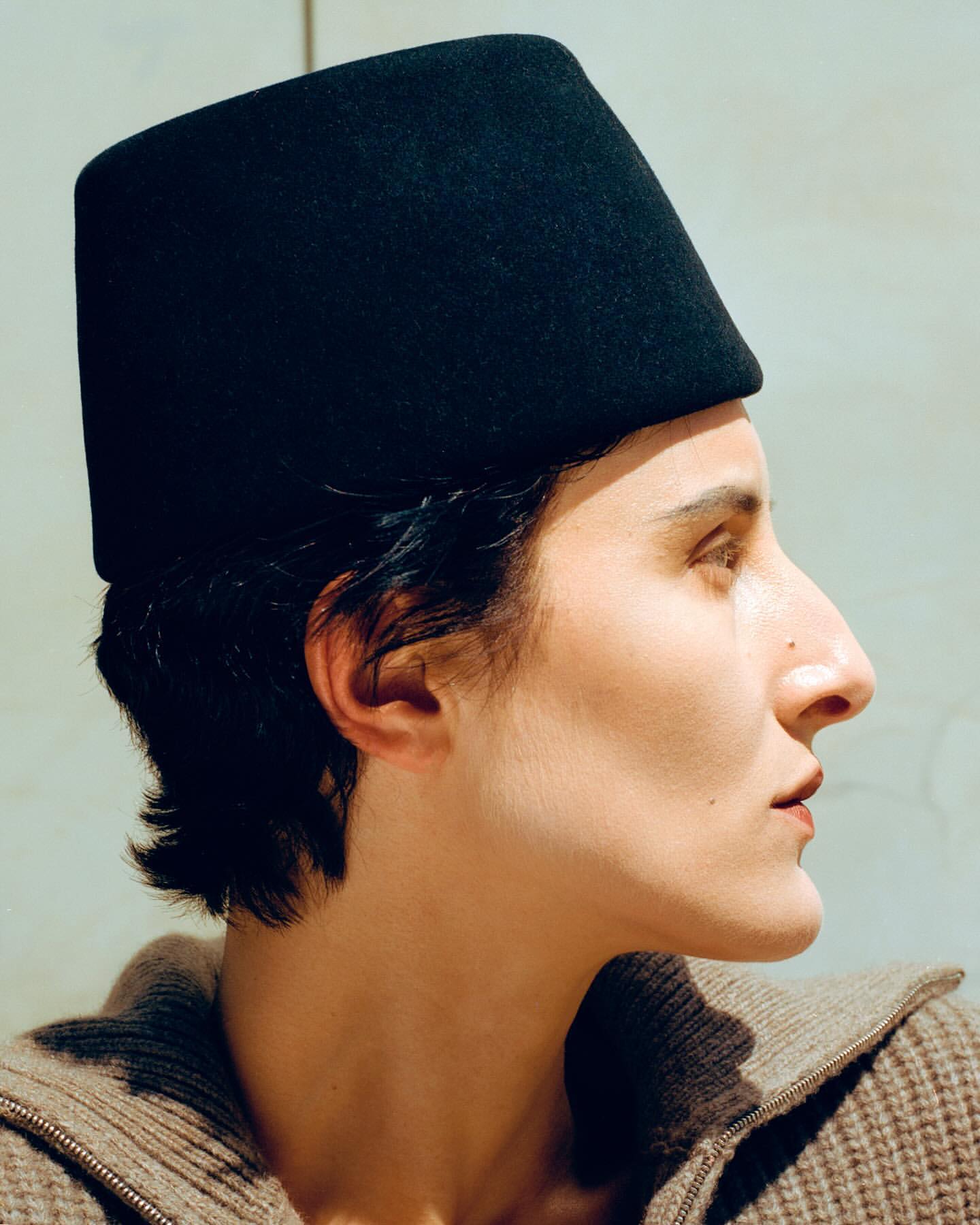 Close-up profile of a woman wearing a black fez-like hat, a tan knitted zip-up top, set against a solid light-colored background.