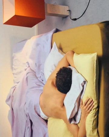 Overhead shot of a person sleeping on a bed with yellow and lavender linens and a bright orange bedside lamp