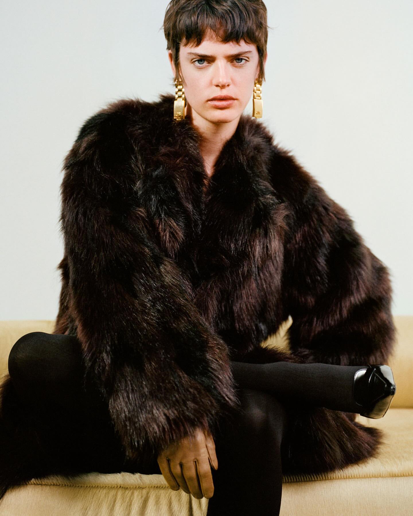Model with short dark hair wearing a voluminous brown fur coat and gold chain earrings while seated on a sofa.