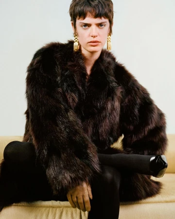 Model with short dark hair wearing a voluminous brown fur coat and gold chain earrings while seated on a sofa.