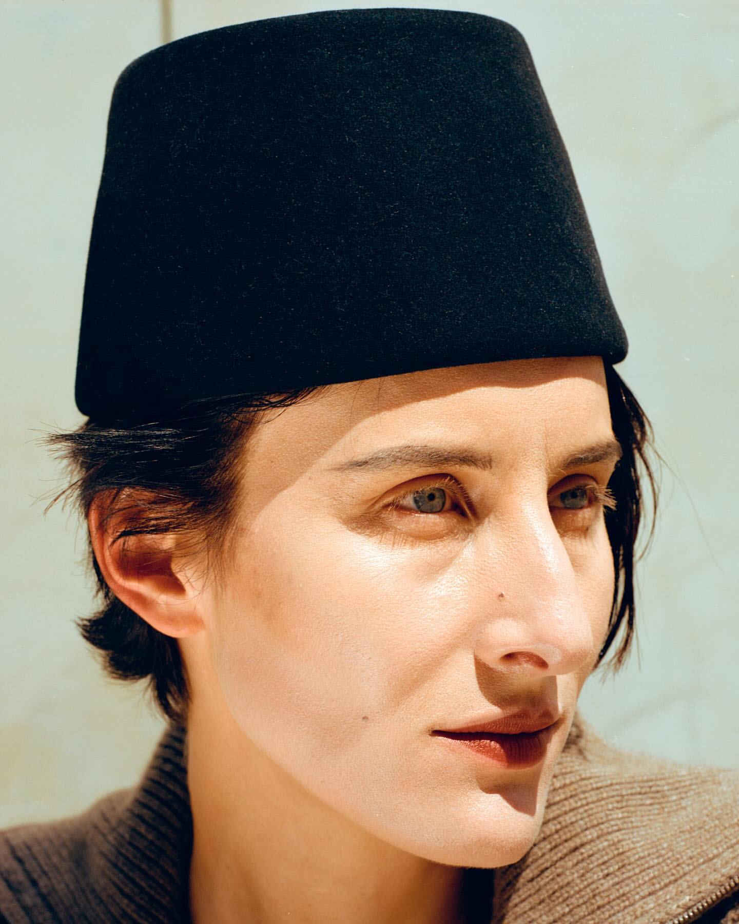 Close-up of a woman with light skin wearing a black fez-like hat and a brown sweater, looking slightly off-center in natural light.