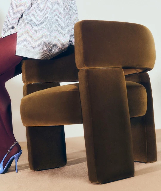 Geometric olive-green armchair with woman's legs in purple stockings and bright blue high heels sitting on it against a neutral backdrop.
