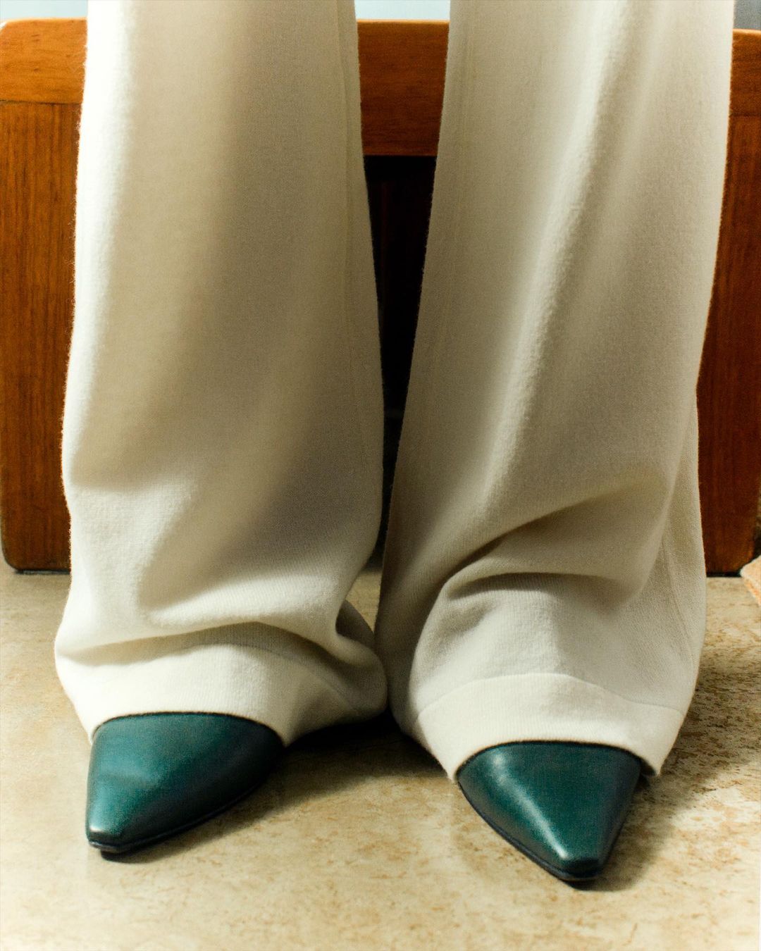 Close-up of light beige trousers over a beige tile floor, combined with a pair of dark teal pointed toe heels and a wooden background.
