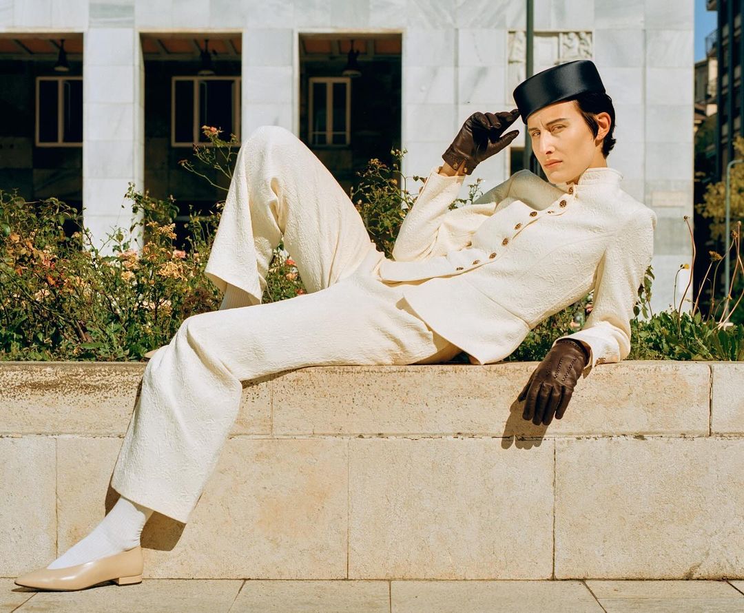 Adult woman poses in a cream-colored suit, hat, and gloves while lying on a stone wall in an urban setting with flowers and a building.