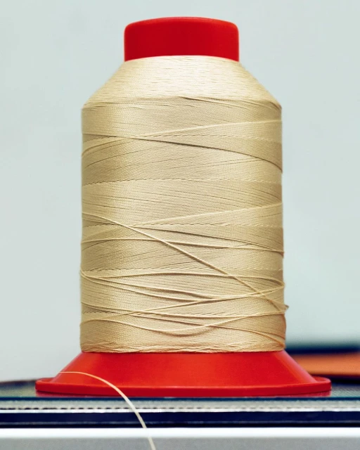 Close-up of a large spool of beige thread with a red plastic base and top, sitting on a surface in a brightly lit indoor space.