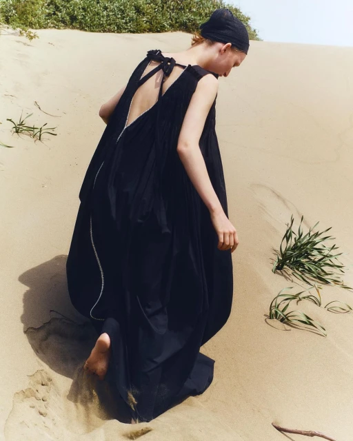 Woman in a dark flowing dress and head covering walking on a sandy beach. Barefoot on sand, facing away, with vegetation visible.
