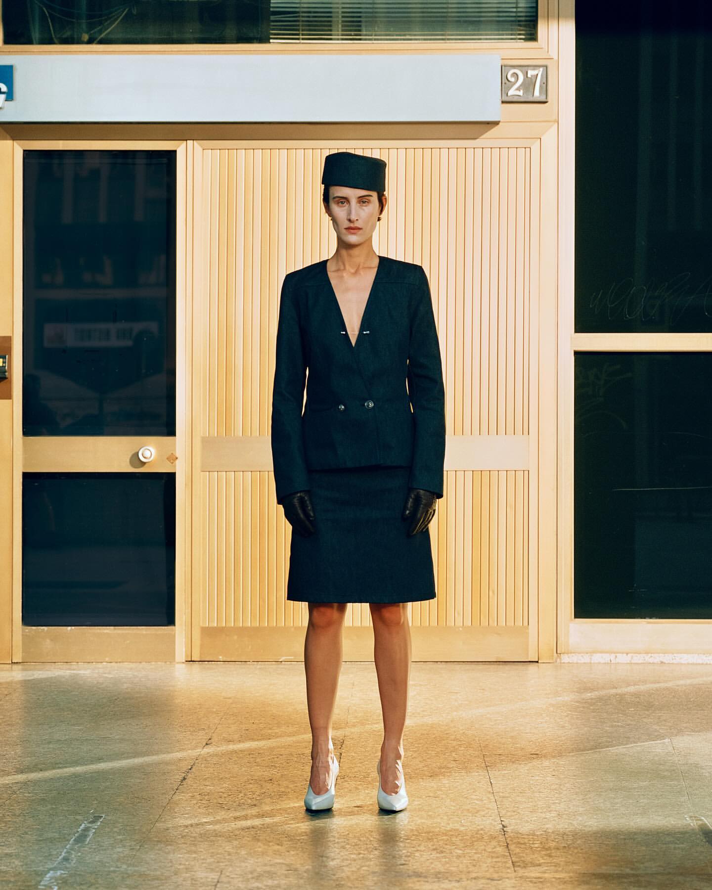 Elegant woman in a dark suit and hat stands in front of a wood-paneled doorway, her white heels adding a contrast to the somber tones.