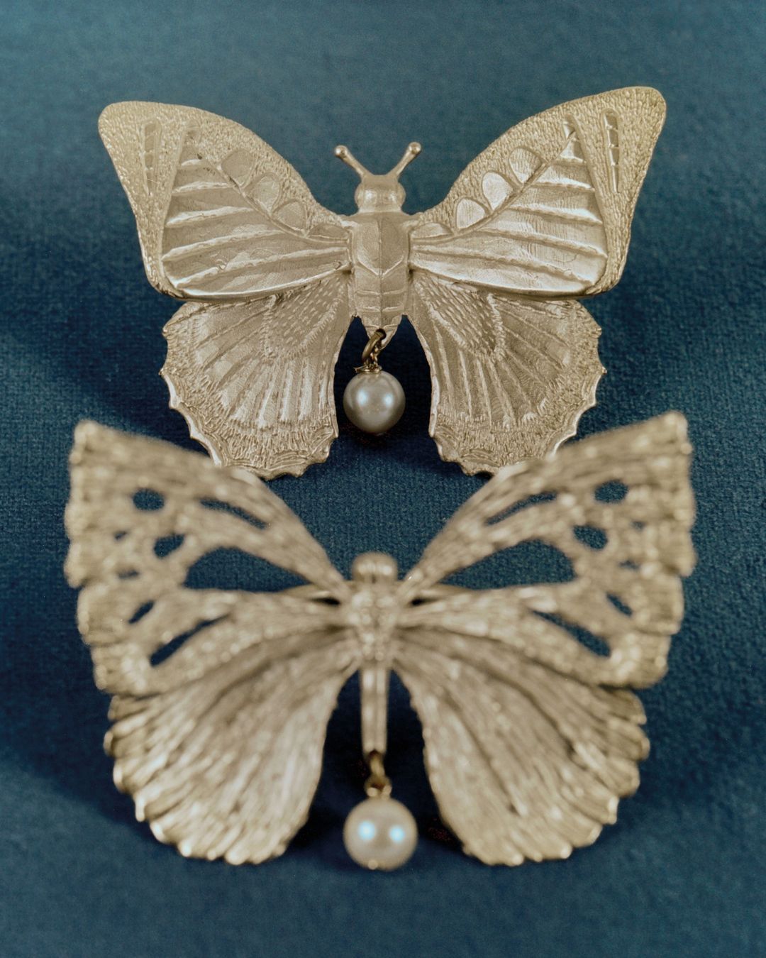 Two elegant butterfly brooches with pearl drops are displayed on a soft teal fabric background in a close-up studio shot.