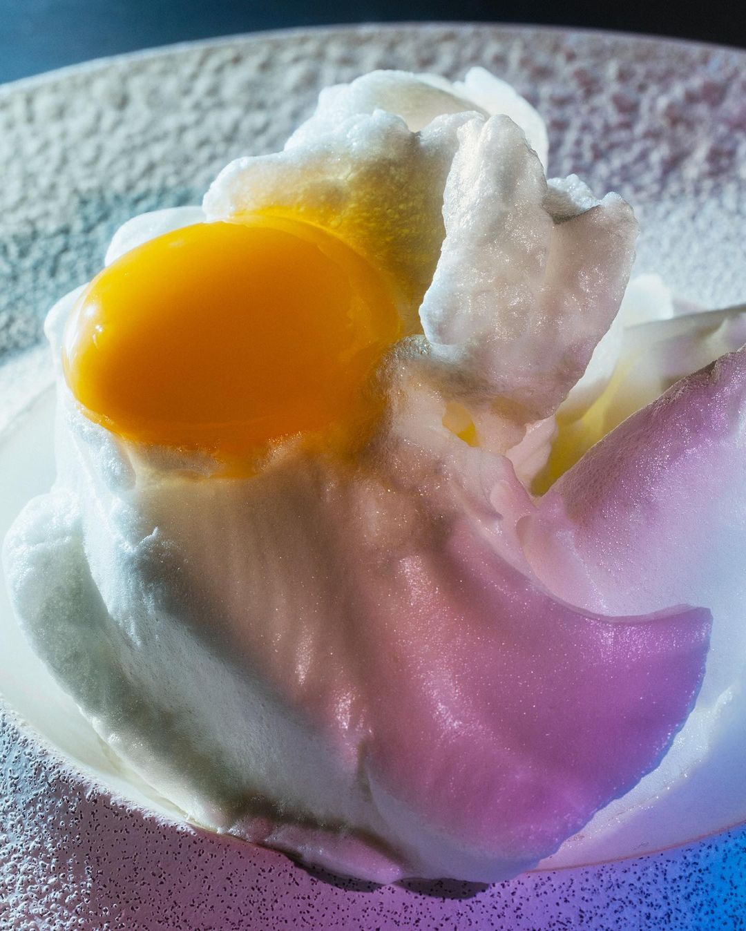 Close-up of an egg cloud on a plate. The fluffy white egg envelopes a bright yellow yolk, presented against a textured background.
