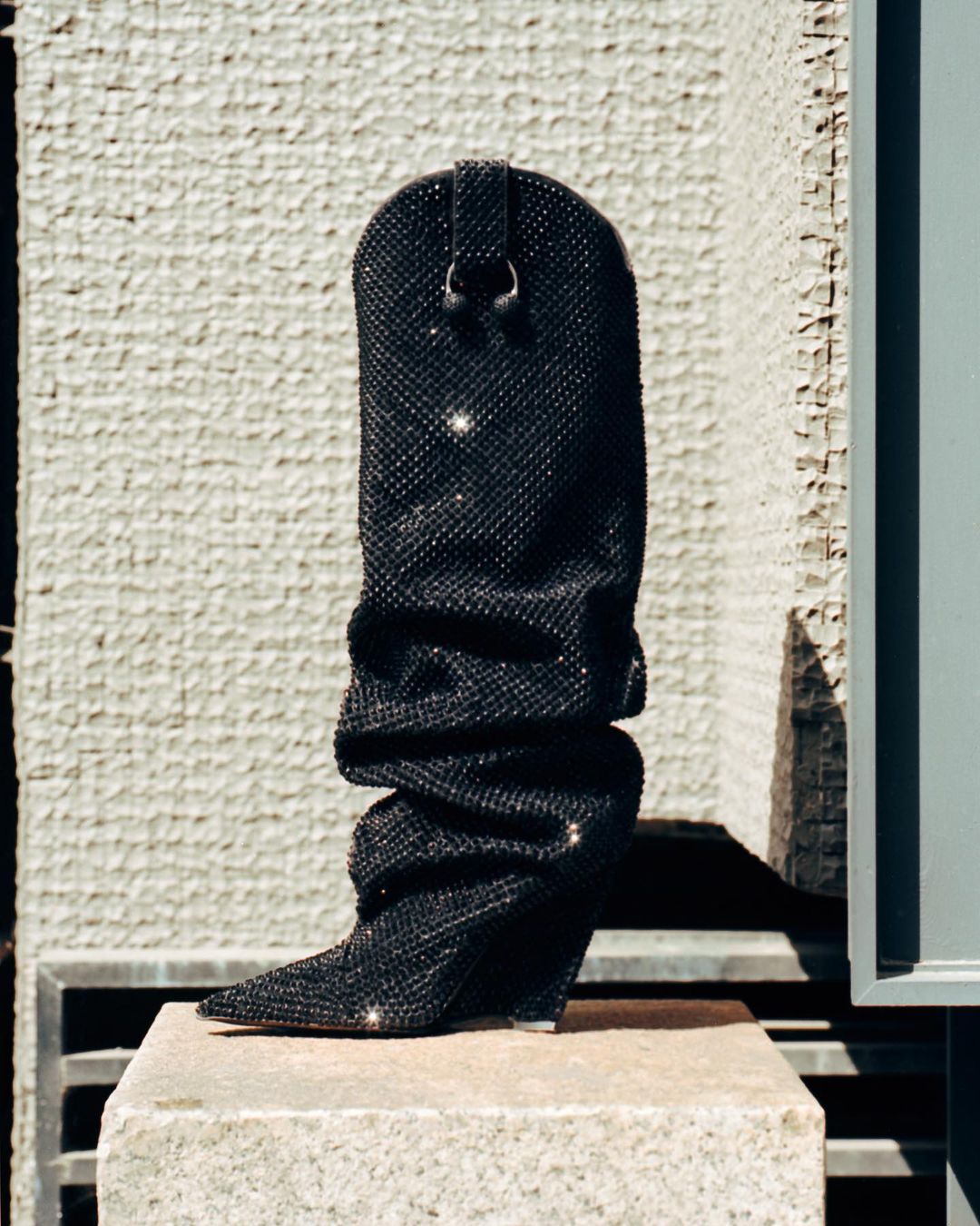 Elegant black cowboy boot covered in rhinestones, showcased on a stone block against a textured concrete wall in bright outdoor lighting.