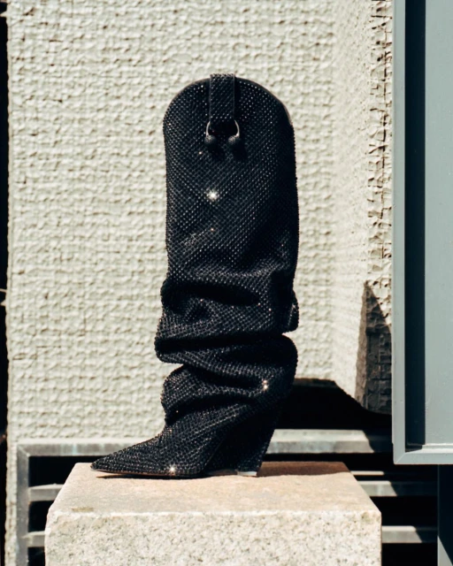 Elegant black cowboy boot covered in rhinestones, showcased on a stone block against a textured concrete wall in bright outdoor lighting.