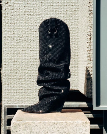 Elegant black cowboy boot covered in rhinestones, showcased on a stone block against a textured concrete wall in bright outdoor lighting.