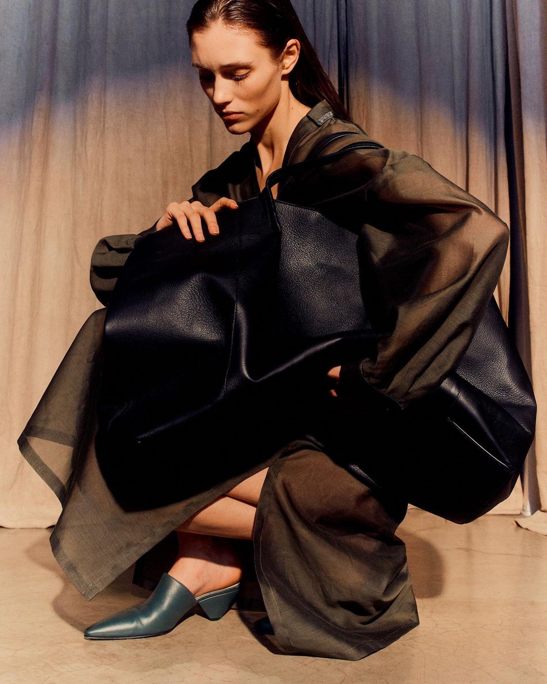 A fashion shot featuring a woman with dark hair crouching and holding a large black tote bag while wearing a green outfit and mules.