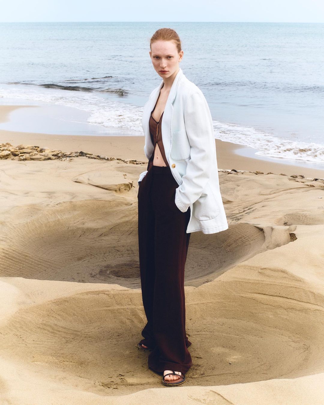 Fashion shot of a redhead adult woman on a sandy beach, wearing a brown top, dark brown pants, and a white jacket against the ocean.