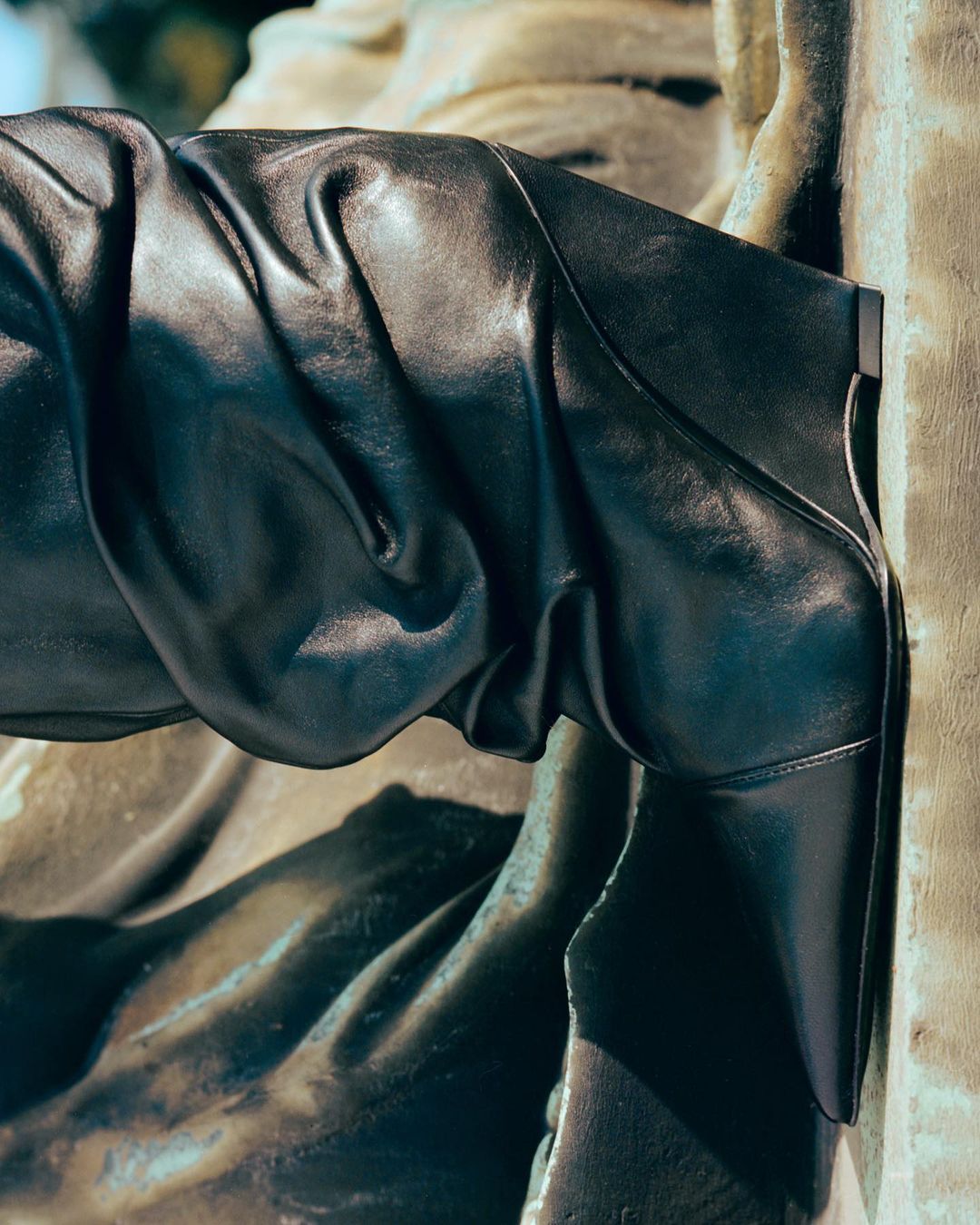 Black leather boot against a textured wall. The boot's design is striking, with sharp lines and a subtle sheen.