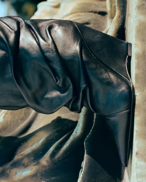 Black leather boot against a textured wall. The boot's design is striking, with sharp lines and a subtle sheen.