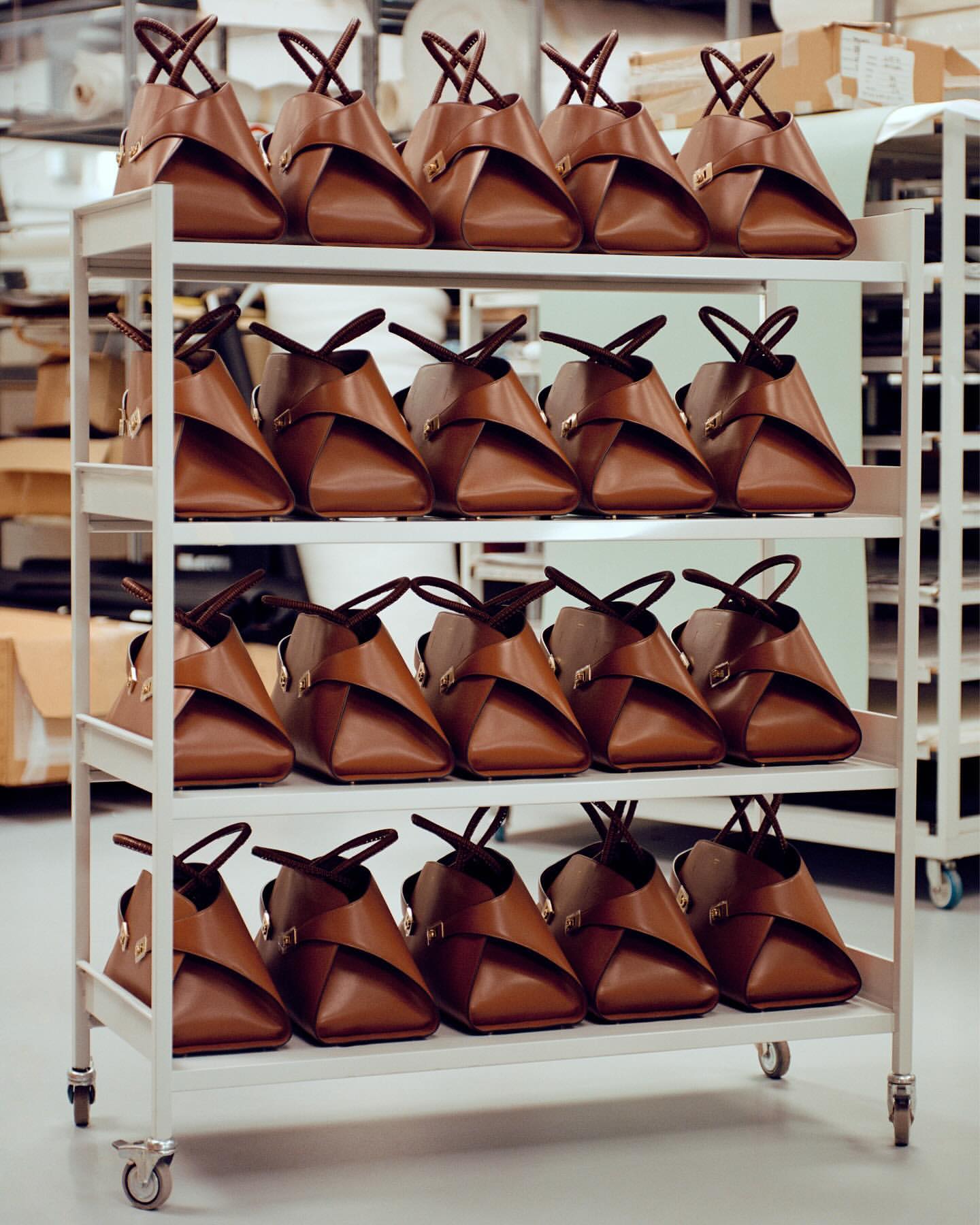 Multiple identical tan leather tote bags with dark brown straps, arranged in rows on a rolling metal rack inside of a building.