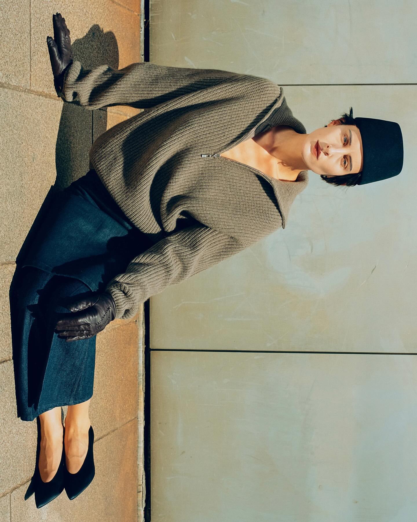 A stylish adult female posing in a studio wearing a black hat, gray sweater, dark blue jeans, black gloves and black high heels.