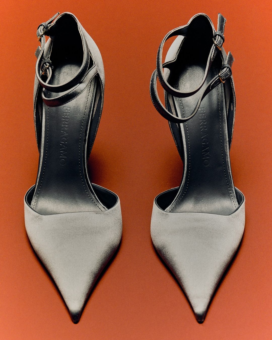 Two grey high-heeled shoes with ankle straps on a bright orange backdrop, studio shot, showcasing fashionable footwear.