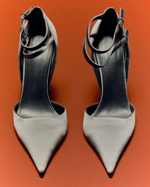 Two grey high-heeled shoes with ankle straps on a bright orange backdrop, studio shot, showcasing fashionable footwear.